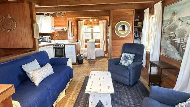 A cozy cabin interior with a blue sofa, a small dining table, a chair, and a kitchen area. - The Back House:  Quaint two bedroom Ocracoke Island cottage (Ocracoke)