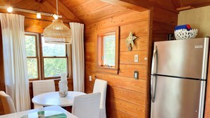 A cozy cabin-style kitchen with wooden walls, a round dining table, white chairs, a stainless steel refrigerator, and a hanging light fixture. - The Back House:  Quaint two bedroom Ocracoke Island cottage (Ocracoke)
