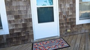 A house with a wooden exterior, a white door with a window, and a welcome mat on the porch. - Hydeaway: Convenient to Restaurants, Shops and Silver Lake (Ocracoke)