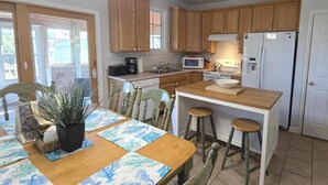 A kitchen with wooden cabinets, a microwave, a refrigerator, and a dining area with a table and chairs. - Hydeaway: Convenient to Restaurants, Shops and Silver Lake (Ocracoke)