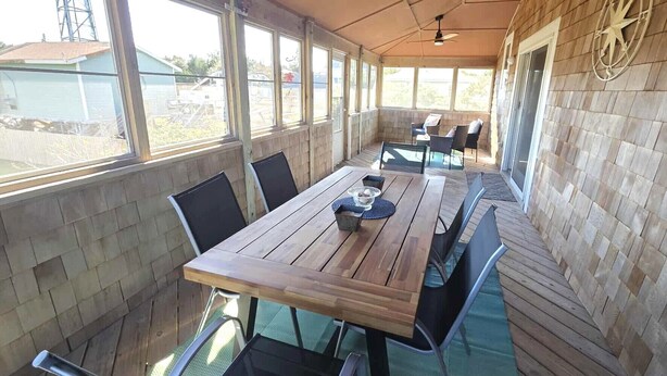 A covered patio with a wooden table and chairs, large windows, and a compass wall decoration. - Hydeaway: Convenient to Restaurants, Shops and Silver Lake (Ocracoke)