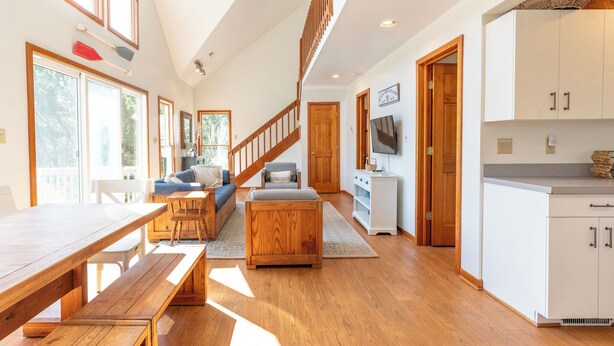 Amplia sala de estar con escalera de madera, una mesa de comedor y una zona de cocina con armarios blancos.