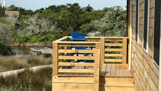 A wooden deck with a blue Adirondack chair, overlooking a body of water and a forested area.