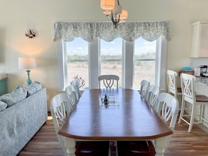 Un coin repas avec une table en bois et des chaises blanches, un canapé à motifs et une vue sur l’extérieur grâce à de grandes fenêtres.