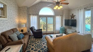 A living room with a sofa, armchair, patterned rug, ceiling fan, and a TV on the wall. - Mermaid Manor: Contemporary spacious home, decks and screened porches (Ocracoke)