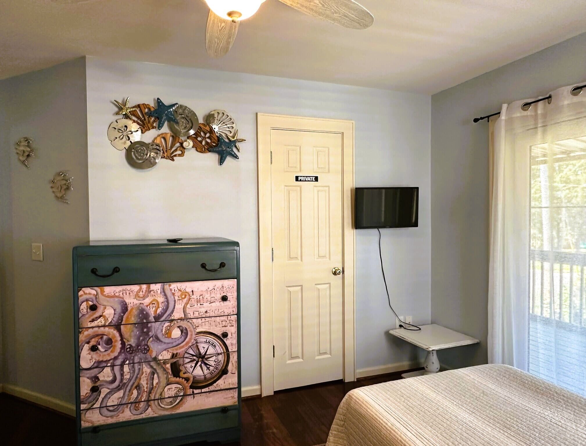 Quarto com cama, penteadeira com detalhes náuticos, porta com a inscrição 'PRIVATE', televisão na parede e janela com cortinas.