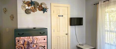 Quarto com cama, penteadeira com detalhes náuticos, porta com a inscrição 'PRIVATE', televisão na parede e janela com cortinas.