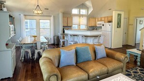 A spacious living room with a leather sofa, dining area with white chairs, a kitchen with white cabinets, and a large window providing natural light. - Mermaid Manor: Contemporary spacious home, decks and screened porches (Ocracoke)