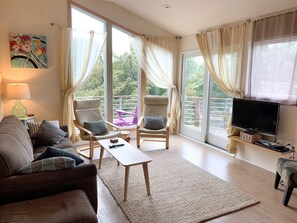 A spacious living room with a sofa, two armchairs, a coffee table, and a TV. Large windows with curtains provide natural light and views of greenery outside. - Friendly Grrrl: 3 bedrooms, 2 Baths. Dog Friendly. (Ocracoke)