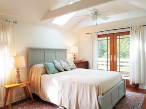 A bedroom with a bed, a nightstand, a lamp, a ceiling fan, and a view of the outdoors through a door. - Cedar Grove Cottage is a lovely pet friendly place to relax and enjoy Ocracoke. (Ocracoke)