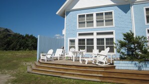 A two-story house with a blue exterior and white trim, featuring a deck with white Adirondack chairs and a small table.