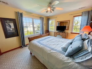 A bedroom with a large bed, a ceiling fan, a dresser, a television, and a window with curtains.