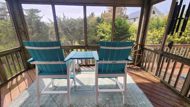 A covered outdoor area with two turquoise Adirondack chairs and a small table, surrounded by a wooden deck and a view of trees and a house. - Nicely Secluded Home, Short Stays Allowed (Ocracoke)