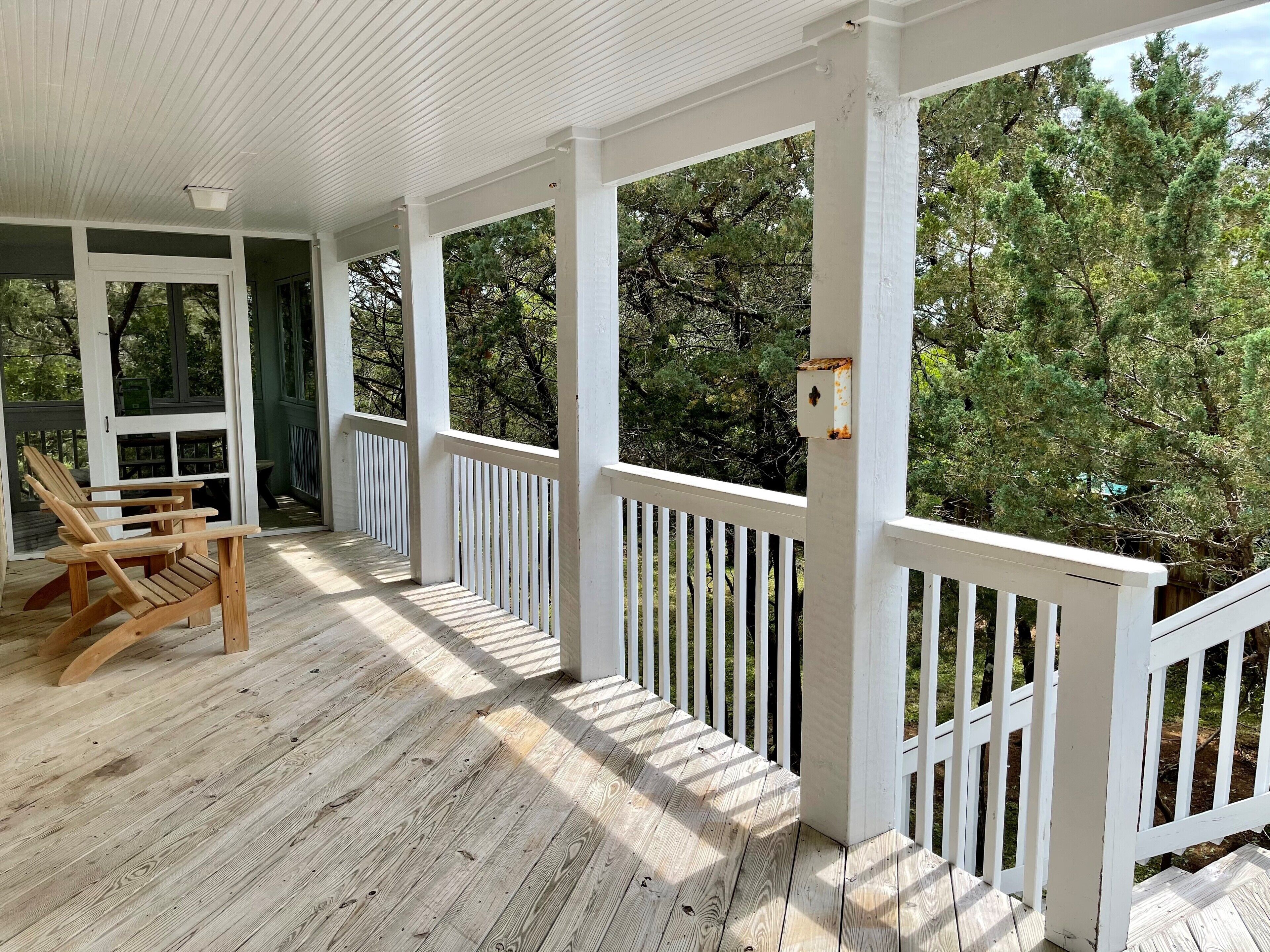 A wooden porch with white railings, a bench, and a view of trees.