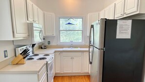 A compact kitchen with white cabinets, a black refrigerator, a stove, and a window with a hanging fish decoration. - Jetts House:  Dog friendly home in private location (Ocracoke)