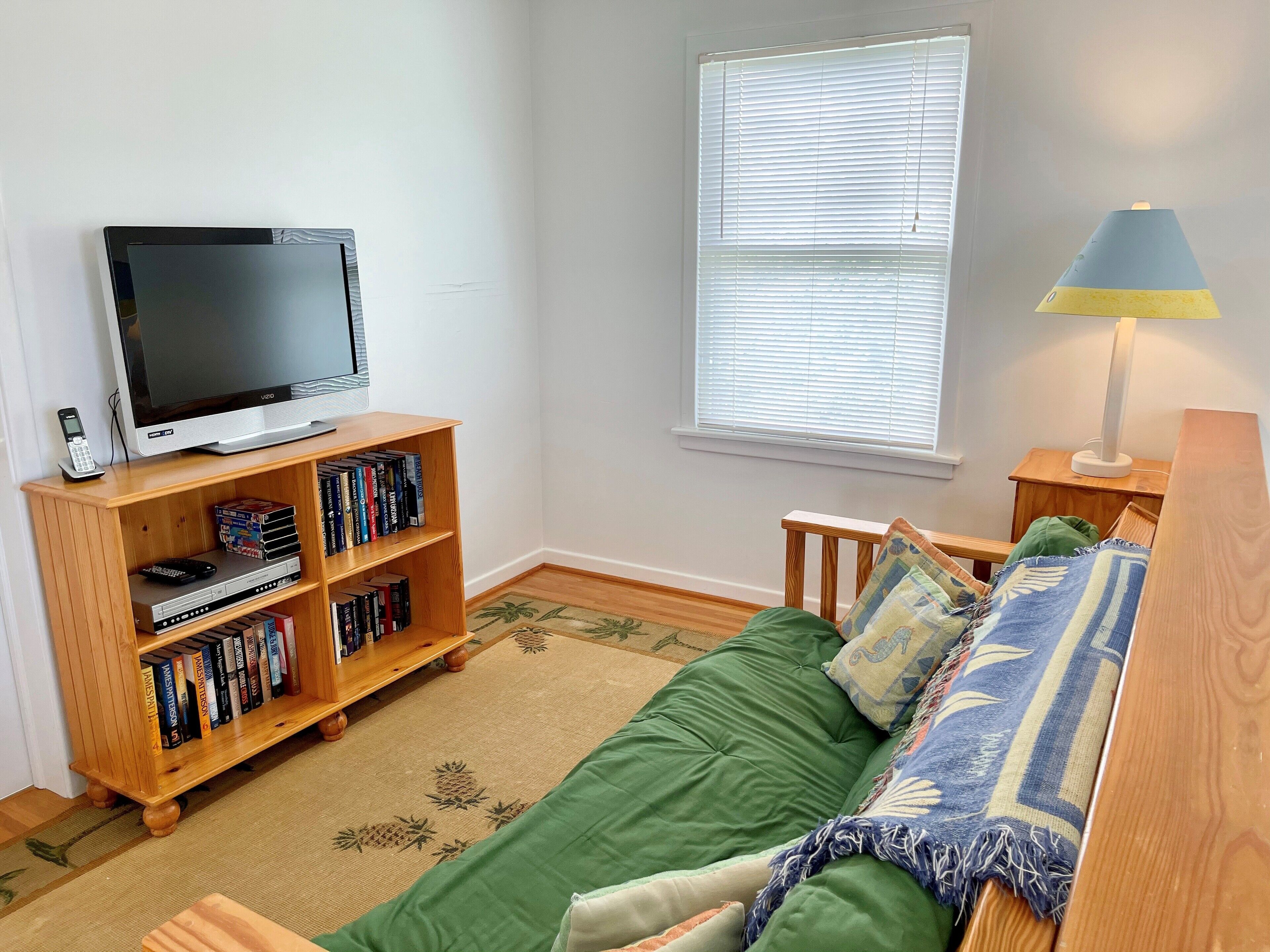 Una habitación acogedora con una televisión sobre un mueble de madera, una estantería, un sofá verde y una ventana con persianas.