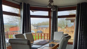 Un coin repas avec une table en bois et six chaises, une vue sur un quartier résidentiel grâce à de grandes fenêtres, et un luminaire au plafond.