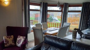 Un salon chaleureux avec un canapé en cuir noir, un fauteuil violet et une table à manger avec des chaises. De grandes fenêtres offrent une vue sur l’extérieur.