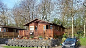 Une cabane en bois, avec des fondations en pierres, une véranda ornée de plantes en pot et une voiture garée devant.