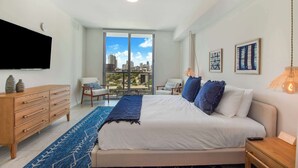 Ein modernes Schlafzimmer mit einem großen Bett, einer Holzkommode, einem Flachbildfernseher und Blick auf die Stadt durch die Fenster.