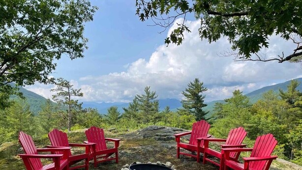 Een groep rode Adirondack-stoelen rond een vuurkorf van natuursteen, in een natuurlijke omgeving met bergen op de achtergrond.