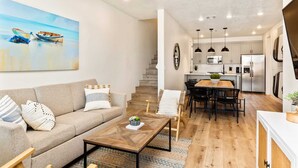 Una sala de estar moderna con sofá, mesa de centro y comedor. Se ve una cocina con electrodomésticos y una escalera al fondo.