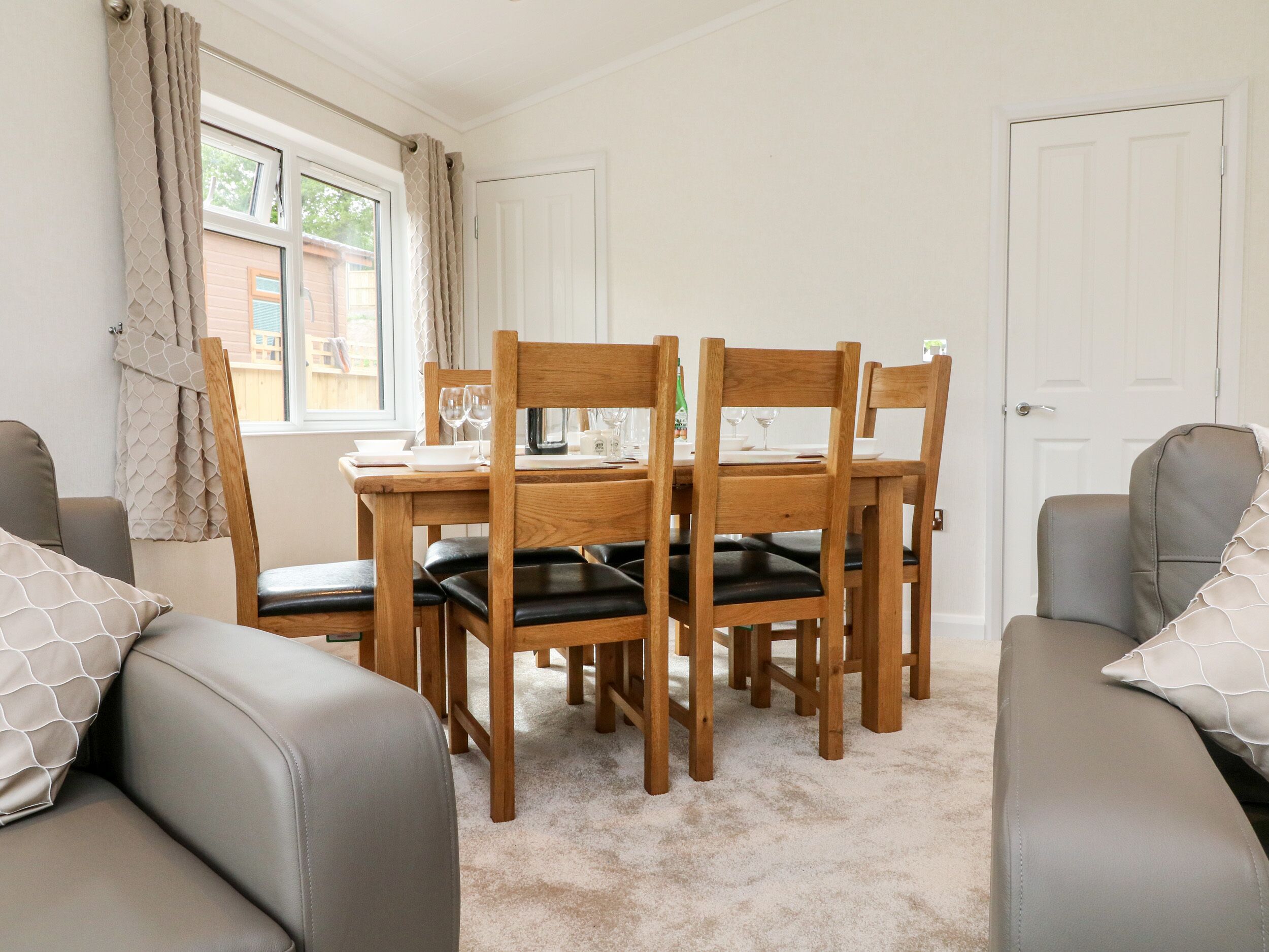 A dining area with a wooden table and chairs, a window with curtains, and a door in the background.