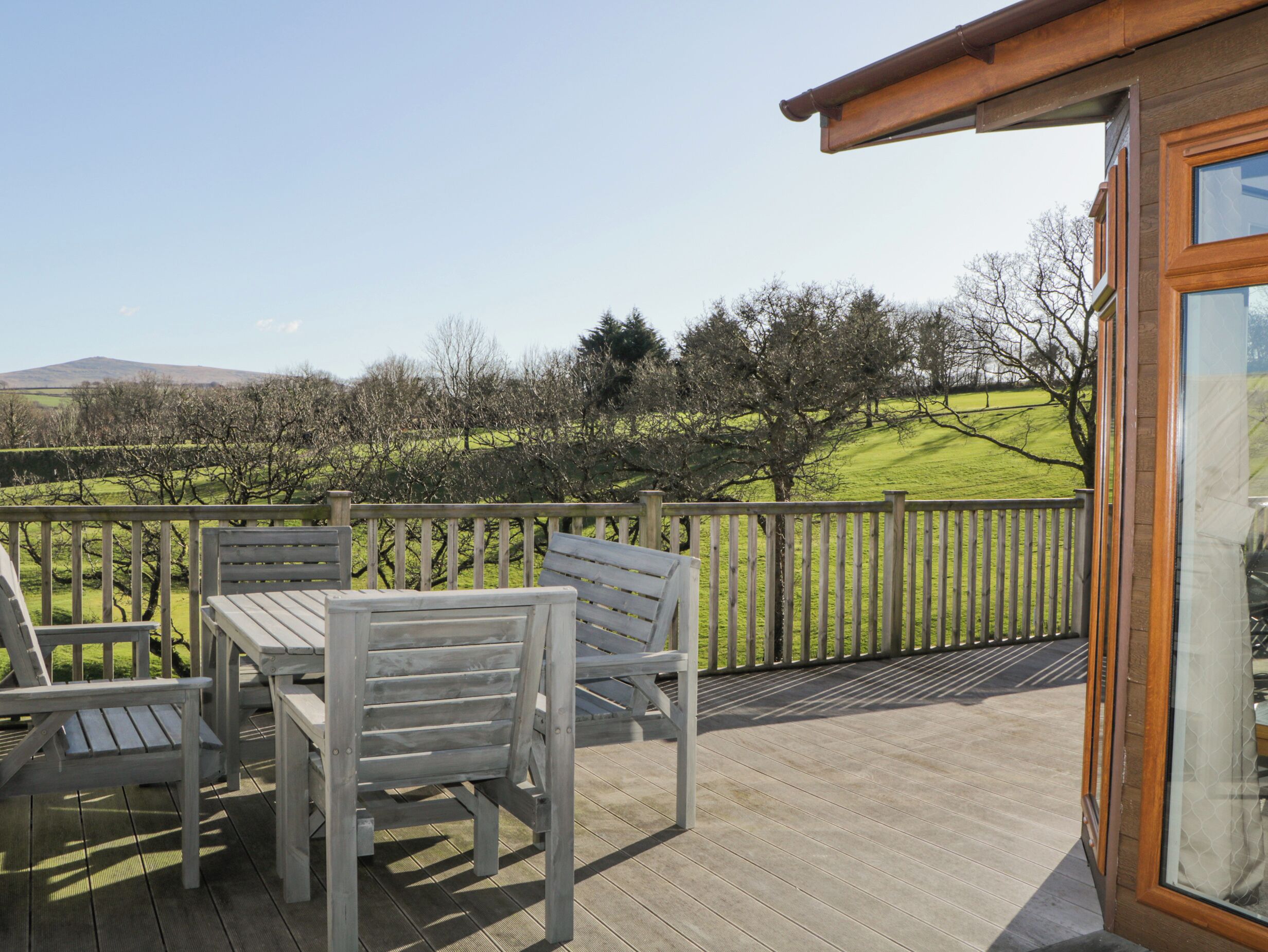Een houten terras met een tafel en stoelen, met uitzicht op een grasrijk landschap met bomen en heuvels.