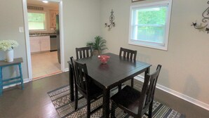 A dining area with a wooden table and chairs, a 'Welcome' sign, a window with blinds, and a kitchen in the background.