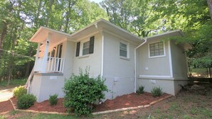 A single-story house with a front porch, a small front yard with mulch and shrubs, and a wooded backdrop.