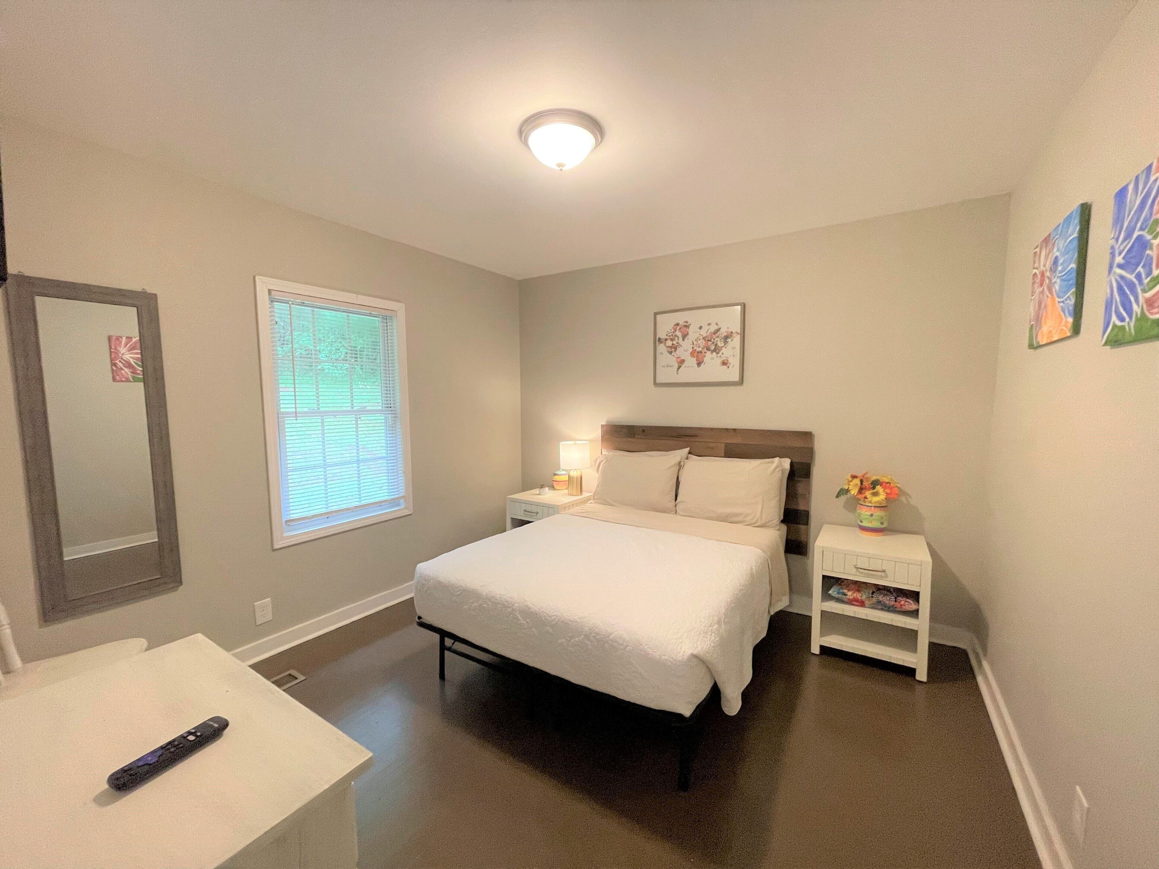 A bedroom with a bed, bedside table, nightstand, window, and a remote control on a table.