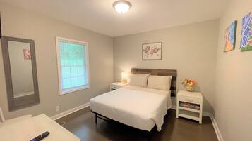 A bedroom with a bed, bedside table, nightstand, window, and a remote control on a table.