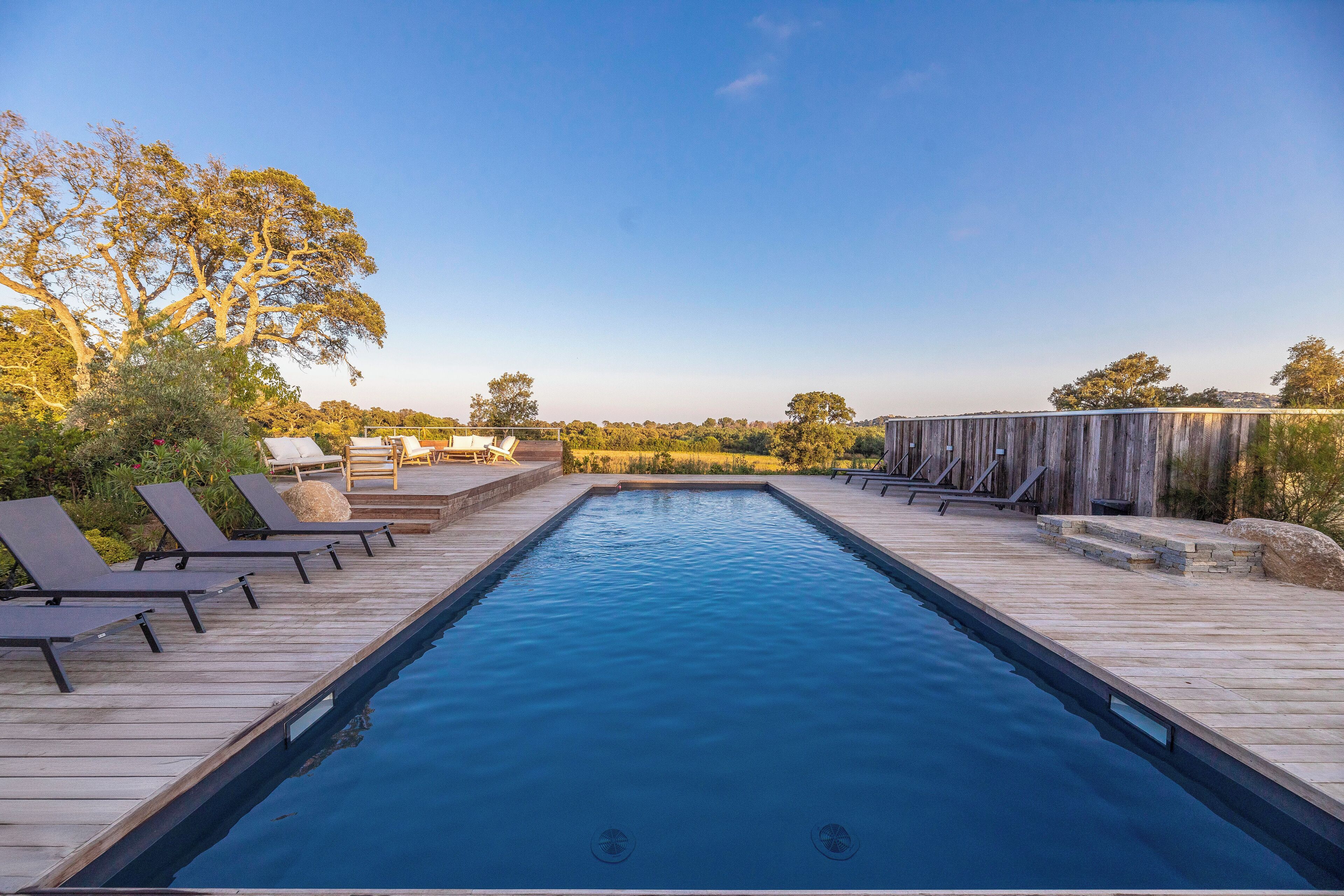 Une piscine rectangulaire dotée d’une terrasse en bois, de chaises longues et d’un espace couvert offrant une vue sur le paysage.