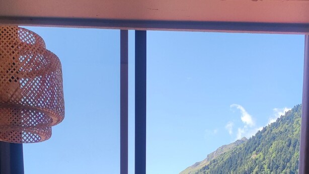 A room with a large window offering a view of a lake and mountains. There is a round table with chairs and a vase on it. - Renovated 4-person studio with lake view Artouste Fabrège (Laruns)