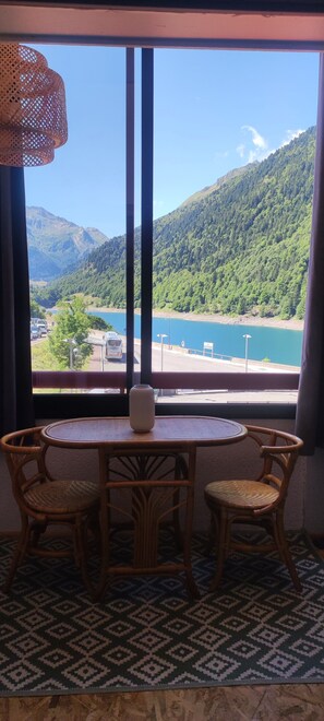 A room with a large window offering a view of a lake and mountains. There is a round table with chairs and a vase on it. - Renovated 4-person studio with lake view Artouste Fabrège (Laruns)
