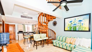 A spacious living area with a wooden staircase, a dining table with chairs, a sofa, and a painting on the wall. - Playa Turquesa D-402 Premier Beachfront Ocean View with 50mbps Wifi (Punta Cana)
