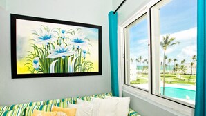A room with a bed, a framed painting of flowers, a window with a view of a pool and palm trees. - Playa Turquesa D-402 Premier Beachfront Ocean View with 50mbps Wifi (Punta Cana)