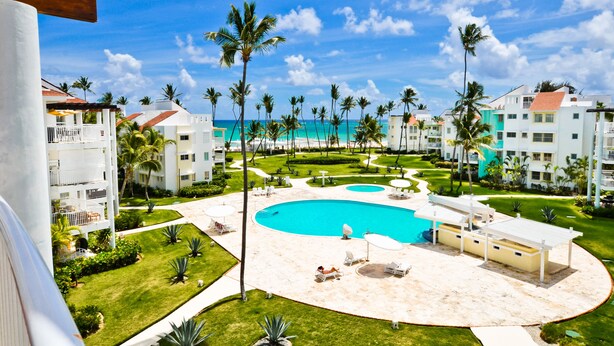 A resort with a central swimming pool, palm trees, and a view of the ocean. - Playa Turquesa D-402 Premier Beachfront Ocean View with 50mbps Wifi (Punta Cana)