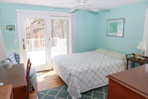 A bedroom with a bed, a desk, a chair, a ceiling fan, and a view through a sliding glass door to a balcony.