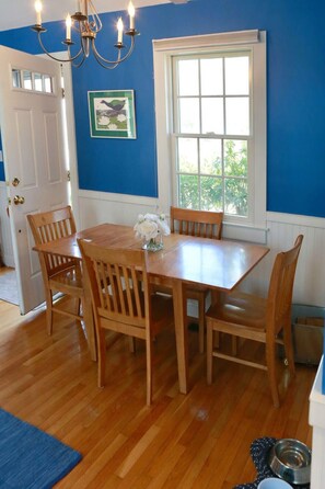 A dining area with a wooden table and chairs, a chandelier, a window with white shutters, and a framed picture on the wall.