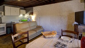 A cozy kitchen with a wooden ceiling, a sofa, a dining table set for two, and a television on a wooden cabinet.