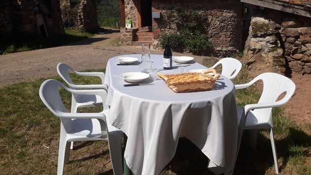 An outdoor dining area with a table set for four, surrounded by white plastic chairs. A basket of bread is on the table, and a bottle of wine is visible. The setting is in front of a stone building with a tiled roof.