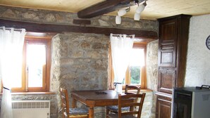 A rustic kitchen with a stone wall, wooden beams, a dining table with chairs, a window with curtains, and a clock on the wall.