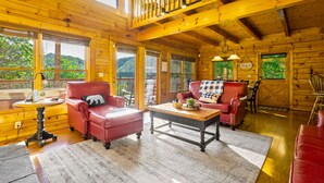 Innenansicht einer Holzhütte mit einem roten Ledersofa, einem Holztisch und Blick ins Grüne.