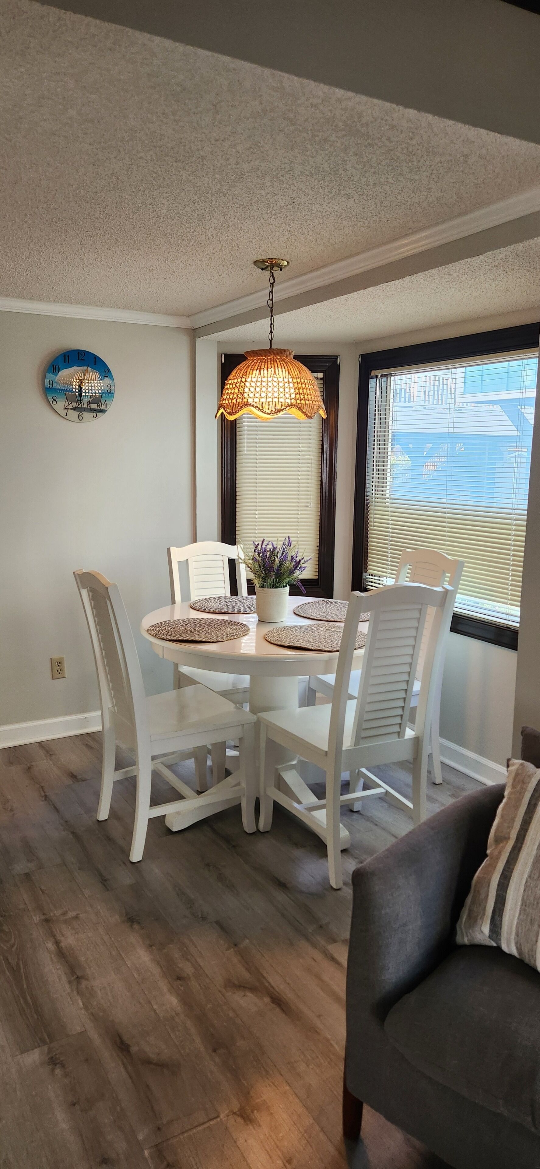 A dining area with a round table, white chairs, a hanging lamp, a clock, and a window with blinds.