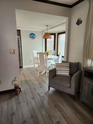 Uma sala de estar com um sofá cinzento, uma mesa de jantar com cadeiras, um candeeiro pendente e um relógio de parede.