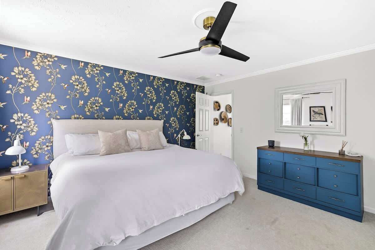 A bedroom with a bed, a ceiling fan, a dresser, and a wall with floral wallpaper.