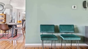 Un comedor con una mesa, sillas y una alfombra. Hay dos sillas verdes junto a una pared con un interruptor de luz y un marco de foto.