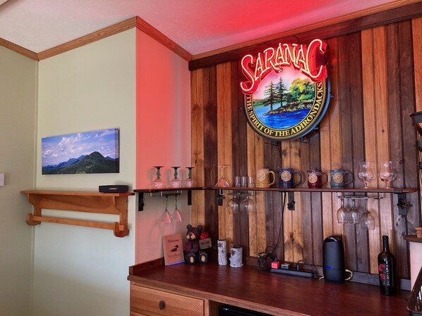 A bar area with a wooden backdrop, a neon sign reading "SARANAC", and various glassware and bottles.
