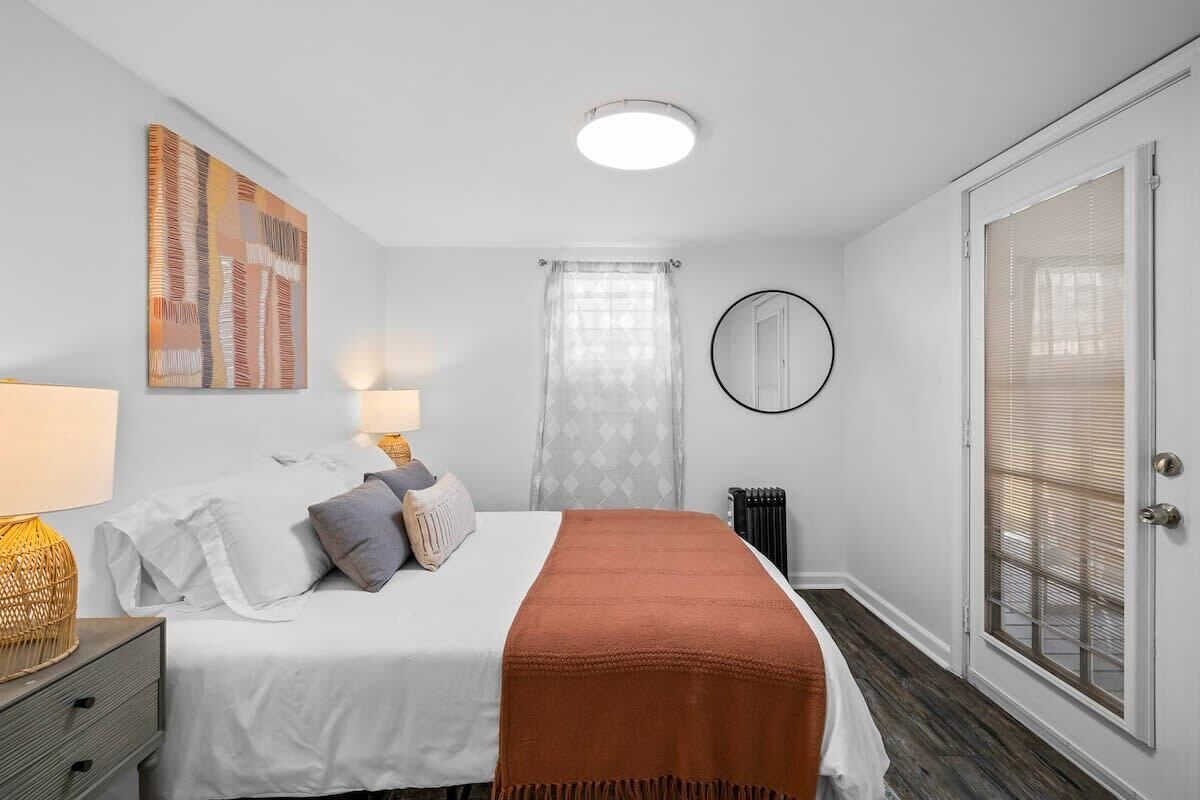 A bedroom with a bed, bedside lamps, a mirror, a window with blinds, and a door.